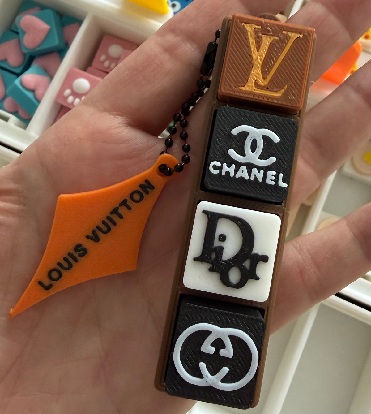 A hand holding a novelty keychain featuring four luxury brand charms: Louis Vuitton, Chanel, Dior, and Gucci, alongside an orange Louis Vuitton tag charm.